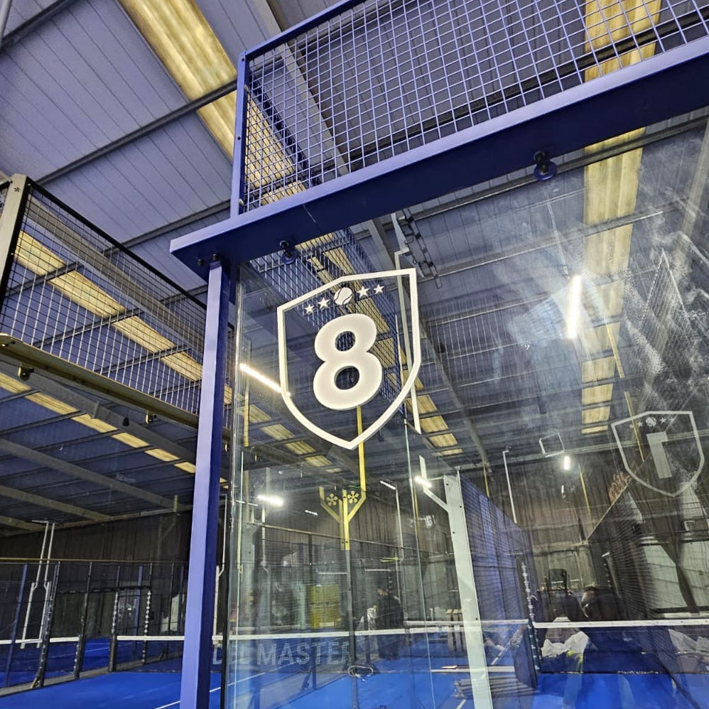 An indoor court with a clear glass wall labeled with a large shield logo and the number 8, overlooking a blue court inside a large industrial-style hall.