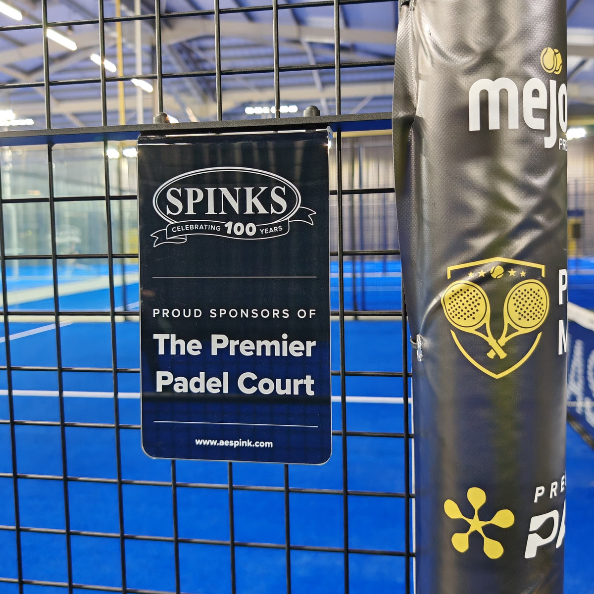 A close-up of a black padded tennis court fence post wrapped with yellow and white branding, including a prominent SPINKS sponsor sign on the mesh fence in front of a blue indoor tennis court.