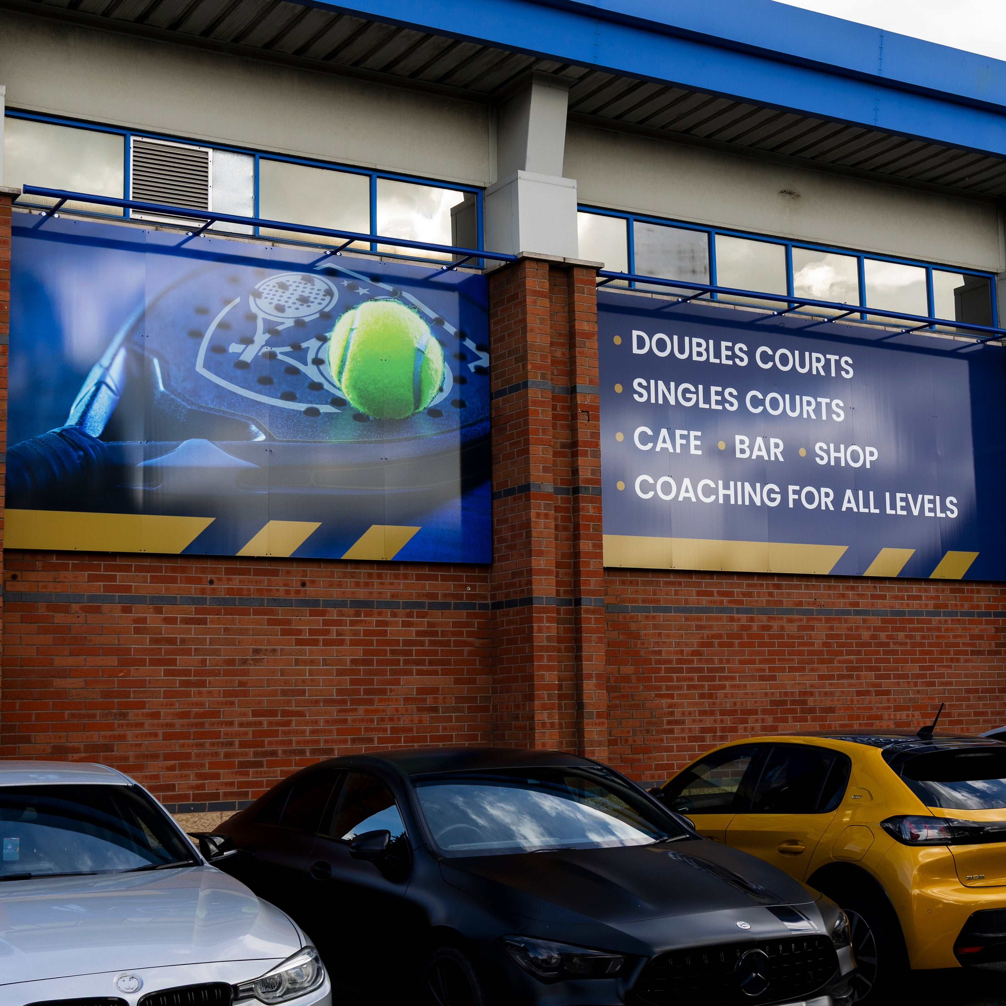 A brick padel club building with large blue banners advertising doubles, singles, cafe, bar, shop, and coaching. P